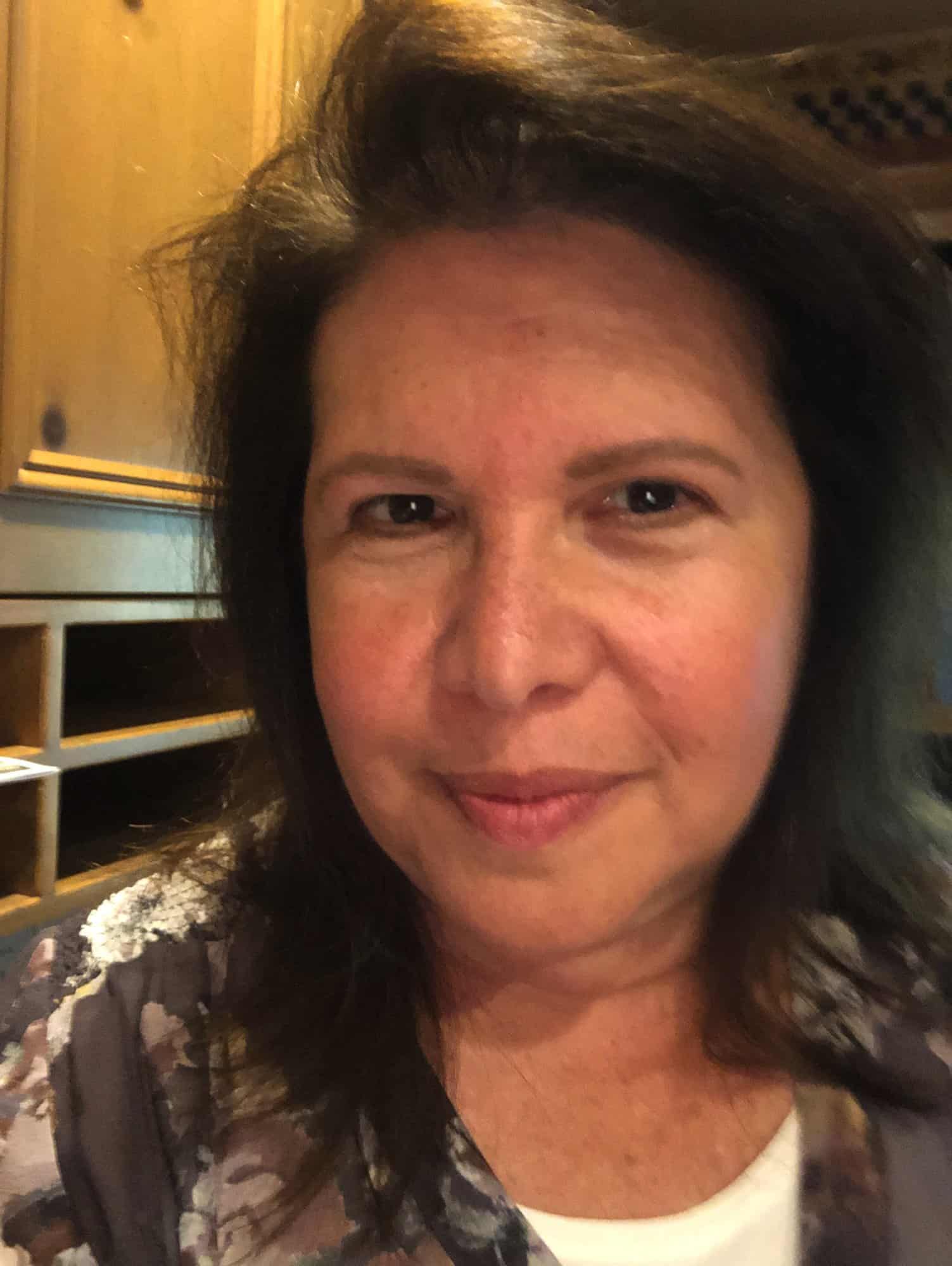A woman with medium-length brown hair and a floral top looks at the camera, standing indoors near wooden cabinets.